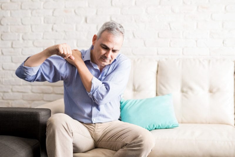 Steroid injection conditions - A middle-aged gentleman sitting on a cream sofa holding his right shoulder with his left hand and rotating his right arm.