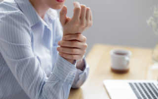 Carpal tunnel syndrome - A female at an office desk wearing a blue and white striped blouse holding her right wrist with her left hand
