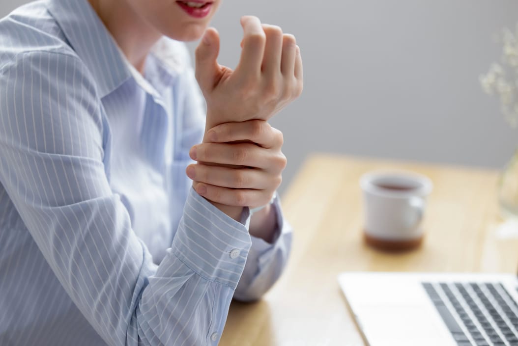 Carpal tunnel syndrome - A female at an office desk wearing a blue and white striped blouse holding her right wrist with her left hand